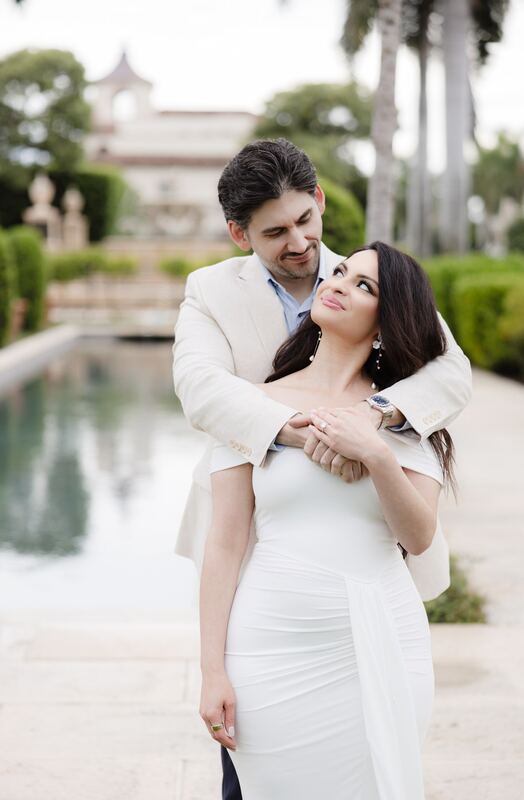Joey wrapping his arms around Iviani from behind beside a palm-lined reflecting pool